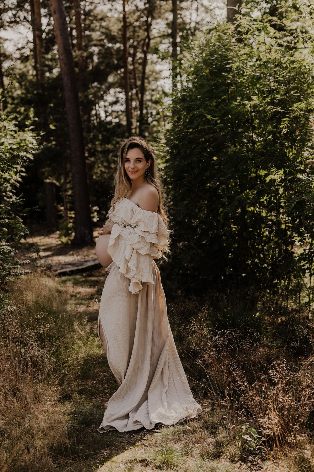 Pregnant woman in an elegant off-shoulder beige ruffled gown posing gracefully in a sunlit forest.