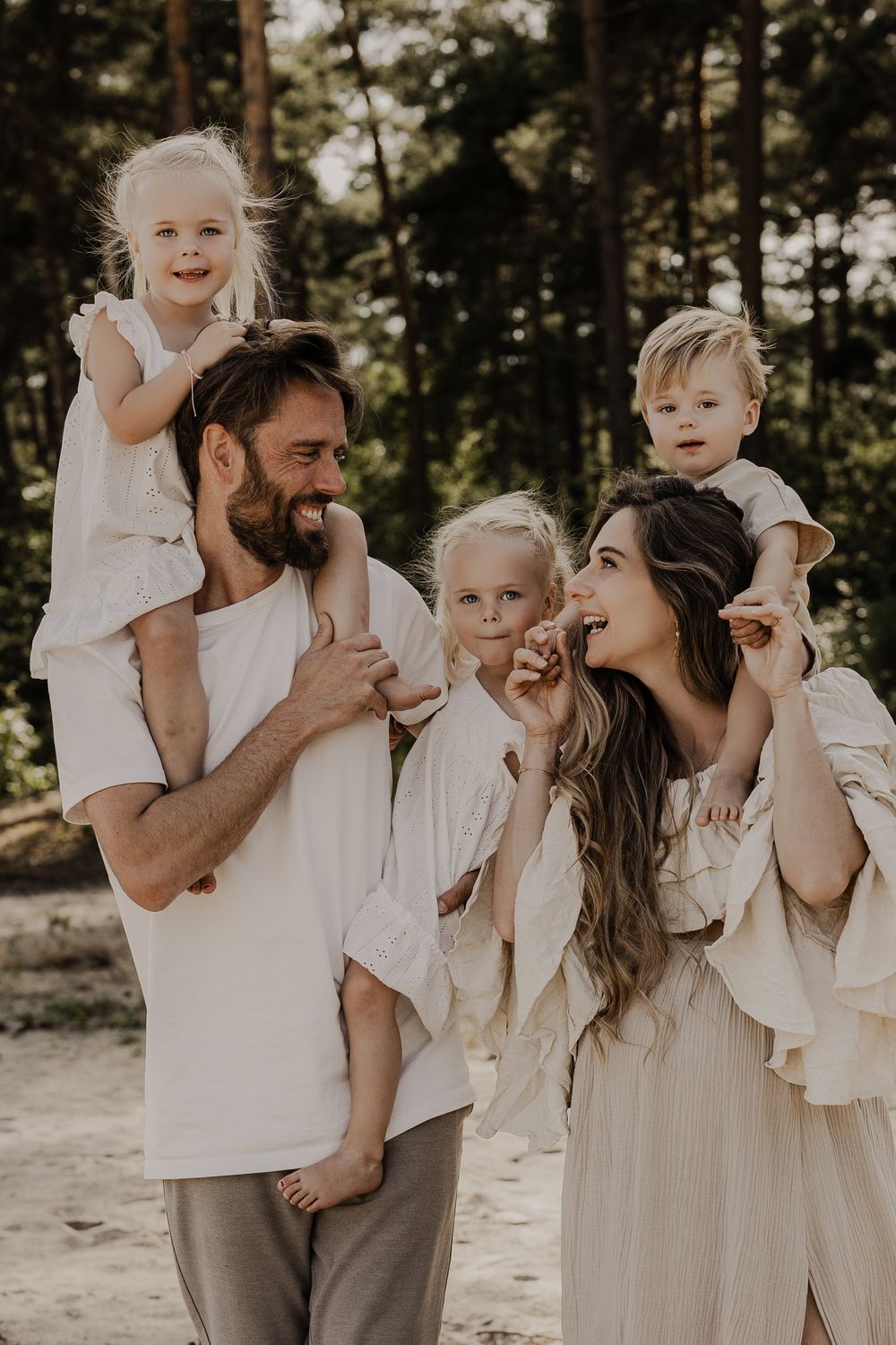 Happy family of five in white and beige outfits with children on shoulders in forest.