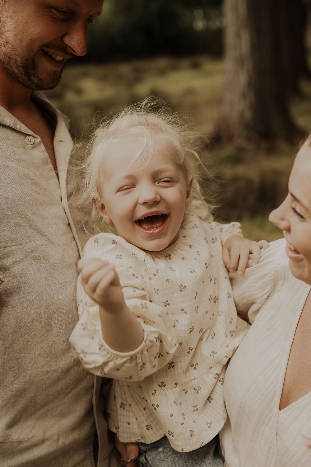 Joyful blonde toddler laughing with delight while being held by her smiling parents outdoors.