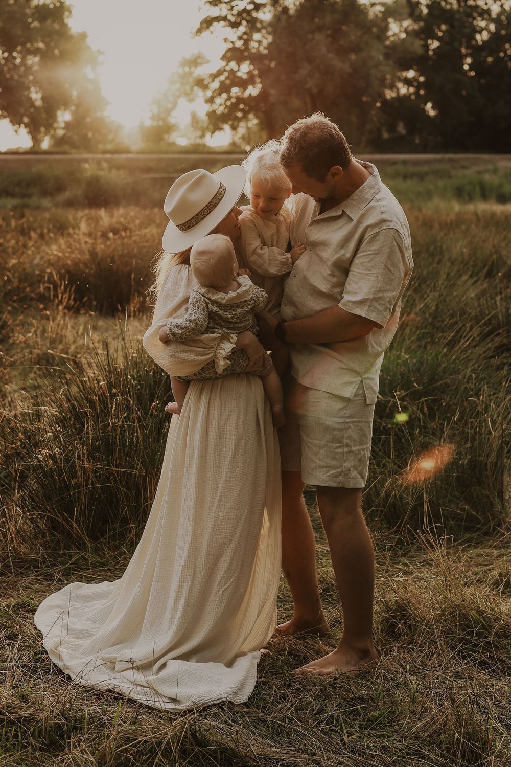 Family of four sharing a tender moment in golden sunset light in a meadow, mother in white dress and hat.