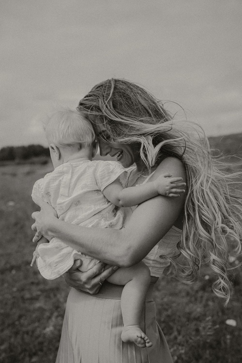 Black and white photo of a mother with long blonde hair holding and embracing her young toddler outdoors.