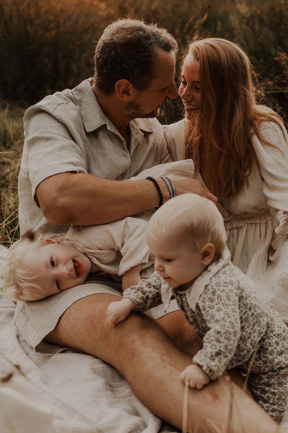Loving couple smiling at each other while two toddlers play on a blanket in a golden meadow.