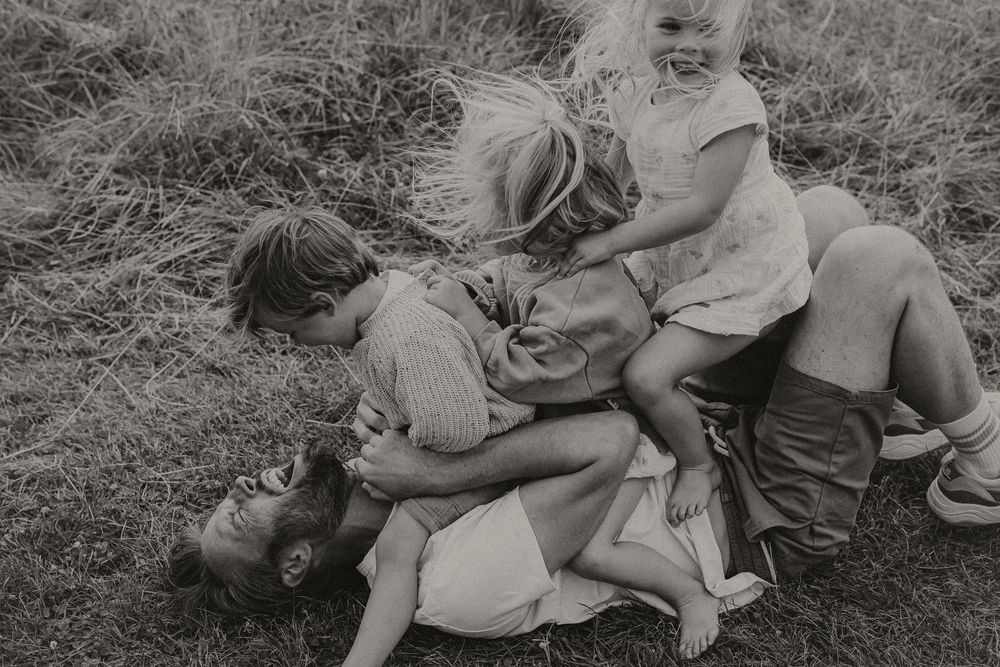 Black and white photo of children playfully piling on an adult lying in a grassy field.