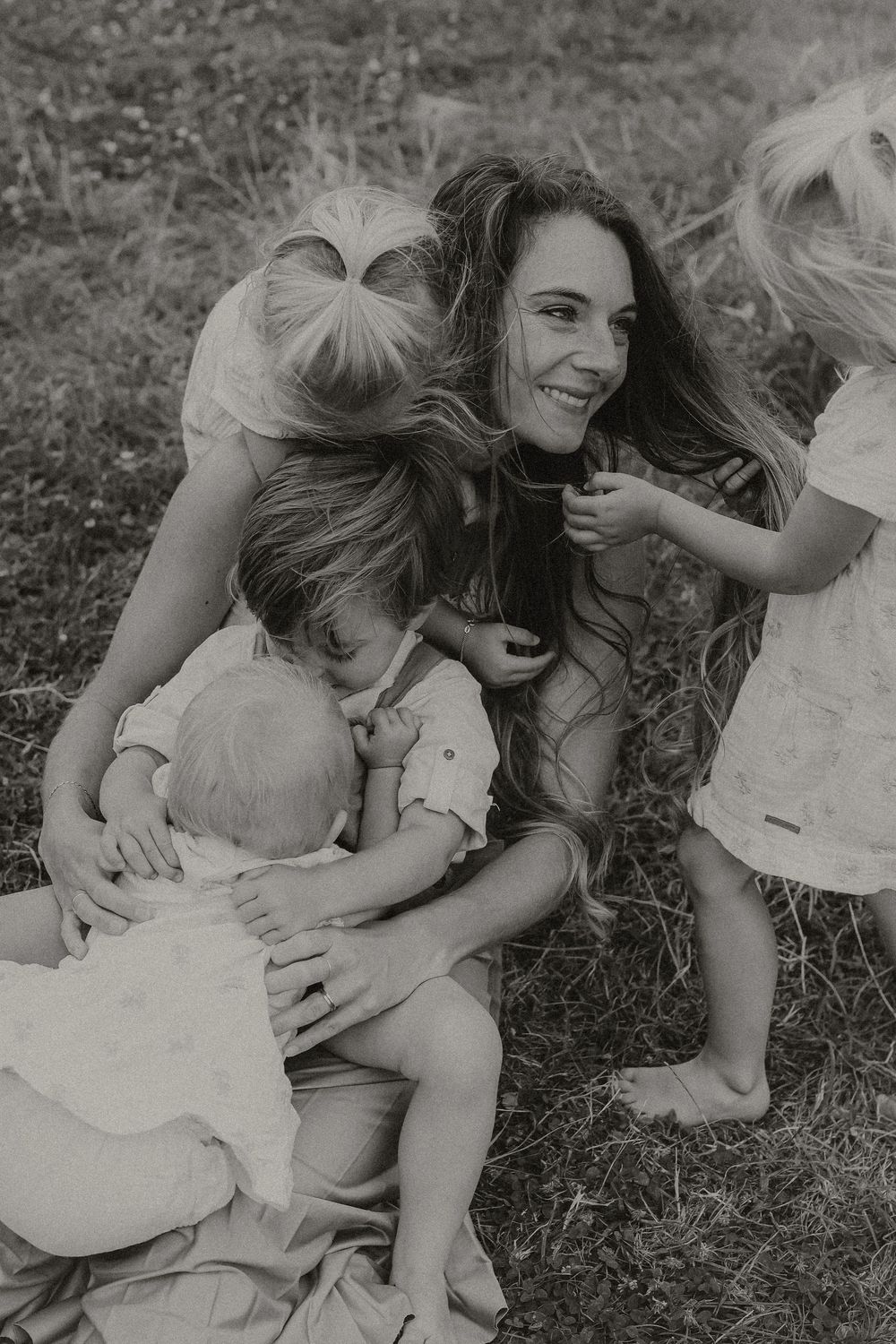 Black and white photo of a smiling mother sitting outdoors on grass, surrounded by three young children hugging her.