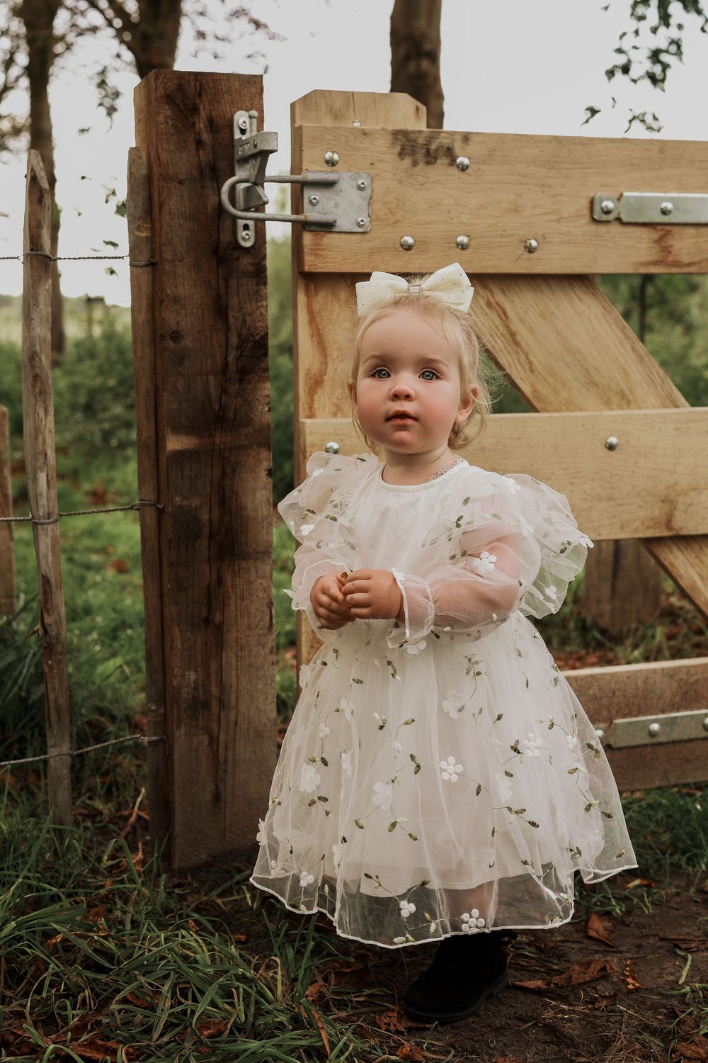 Adorable toddler girl in a white floral tulle dress with a bow standing by a wooden farm gate.