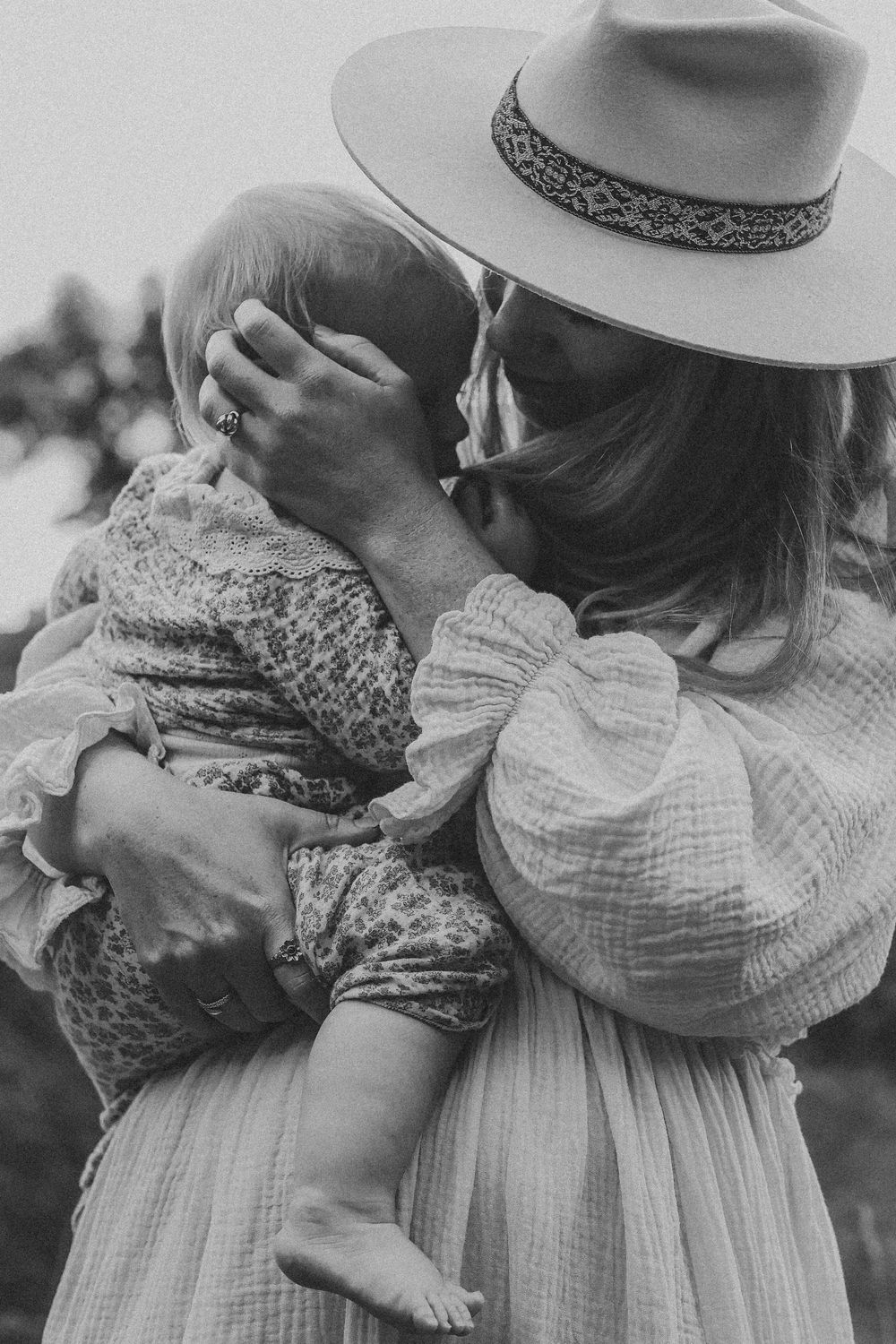 Black and white portrait of a mother in a wide-brim hat tenderly holding her baby close outdoors.