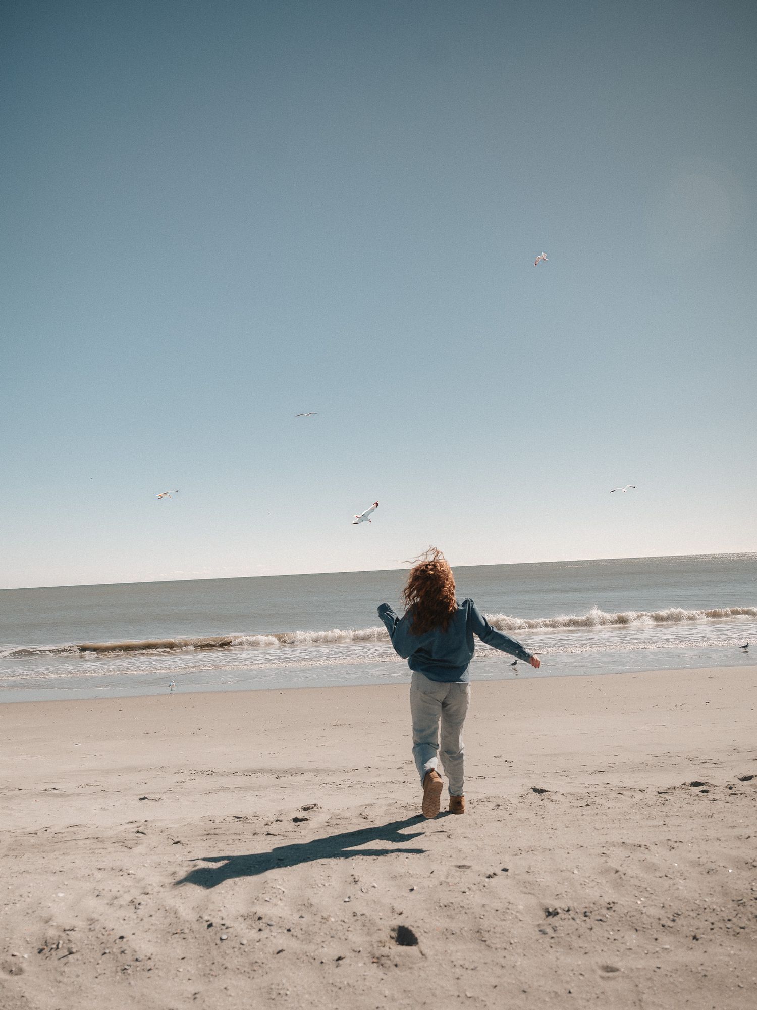 A figure casts a long shadow while walking along a serene beach shoreline at sunset.