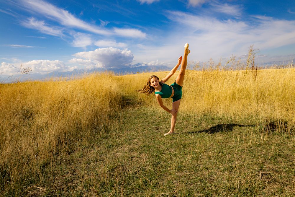 Dance pose with extended leg in green outfit amid golden grass field.