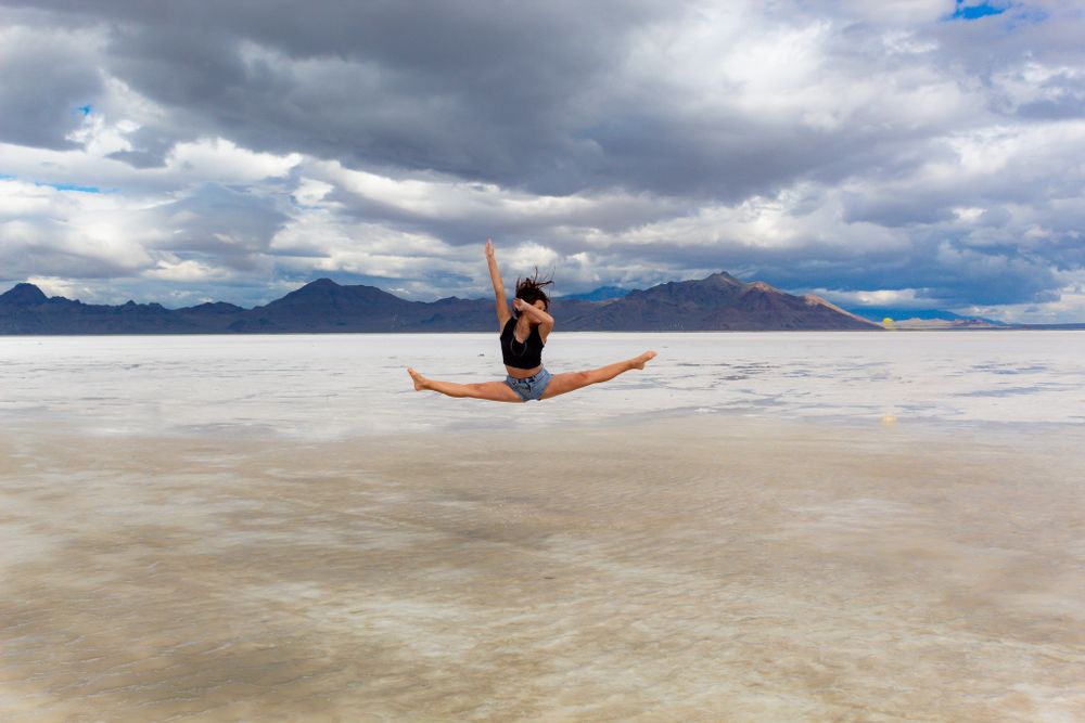 Dancer performs a perfect split jump on a salt flat with dramatic storm clouds and mountains.