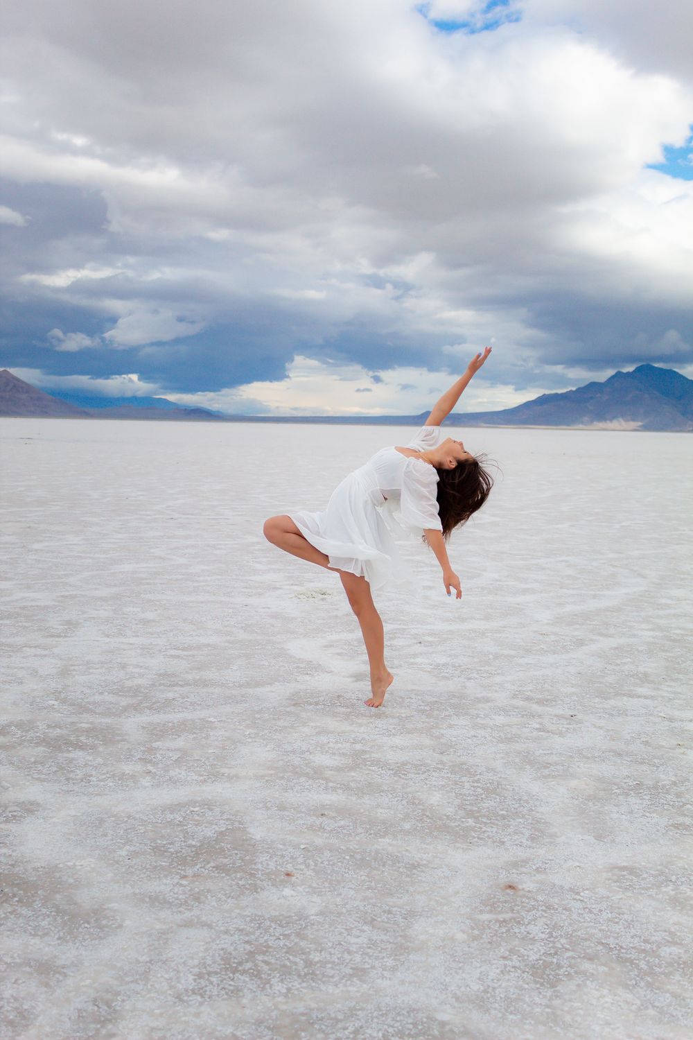 Graceful dance pose in white dress captured on stark salt flats under cloudy skies.