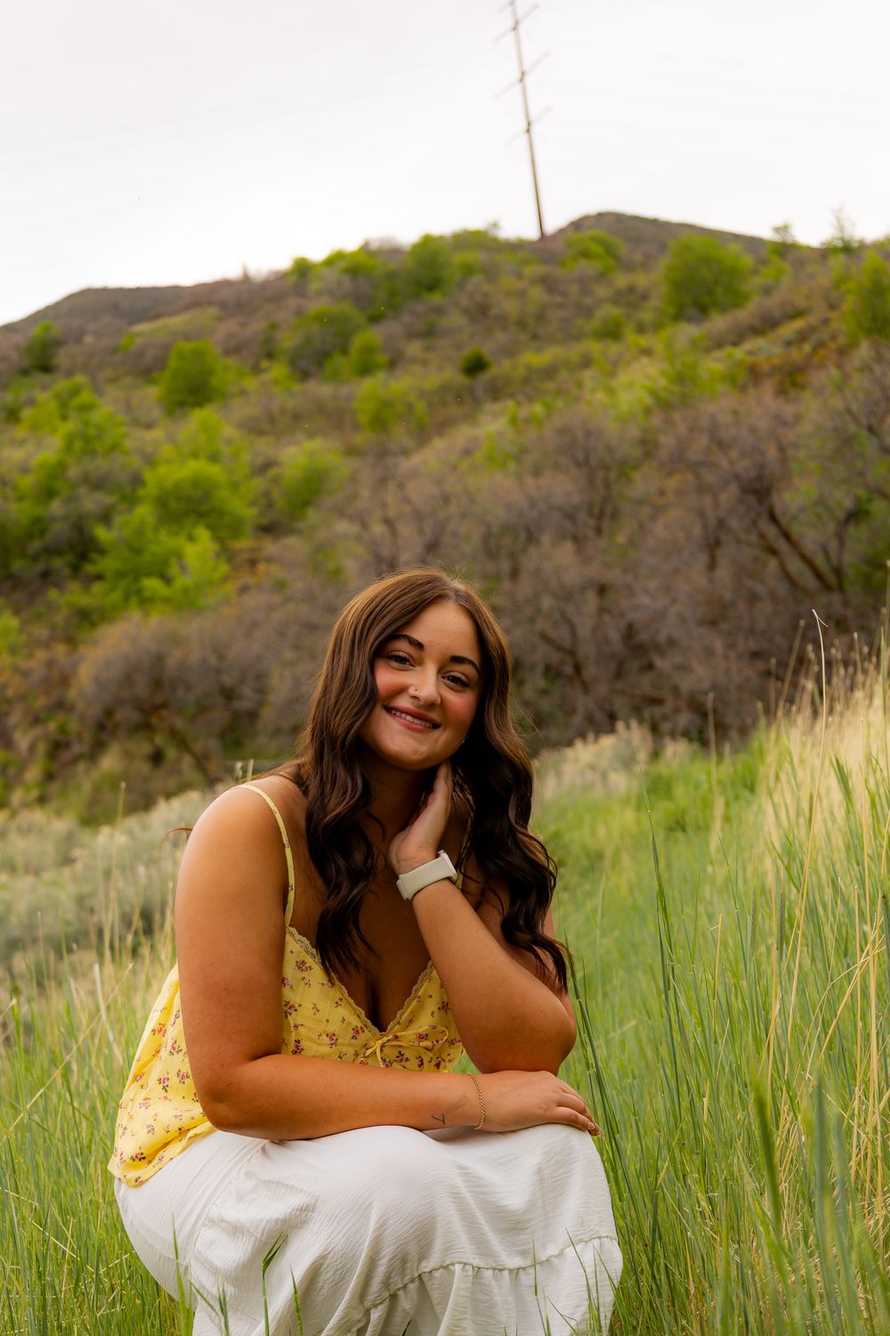 Young woman in yellow top and white skirt sitting in green meadow with tree-covered hills in background.
