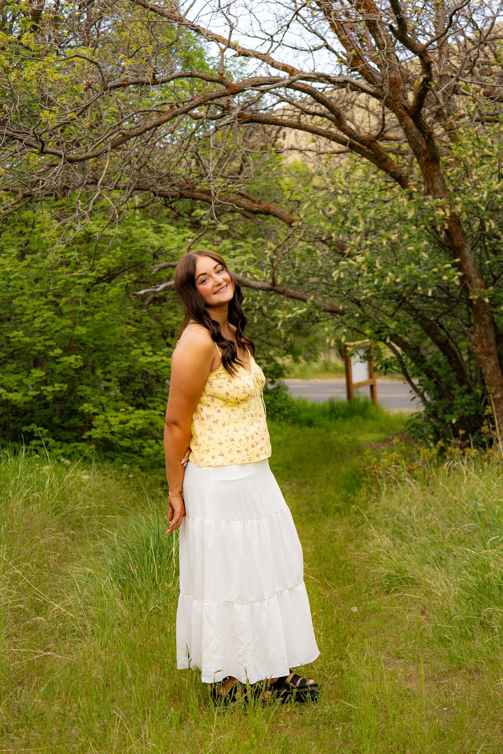 Young woman in yellow floral top and white maxi skirt smiling outdoors among lush green trees and grass.