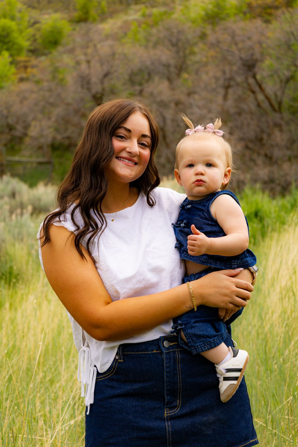 Young woman in white top and denim skirt holds a baby girl in blue outfit in a green meadow.