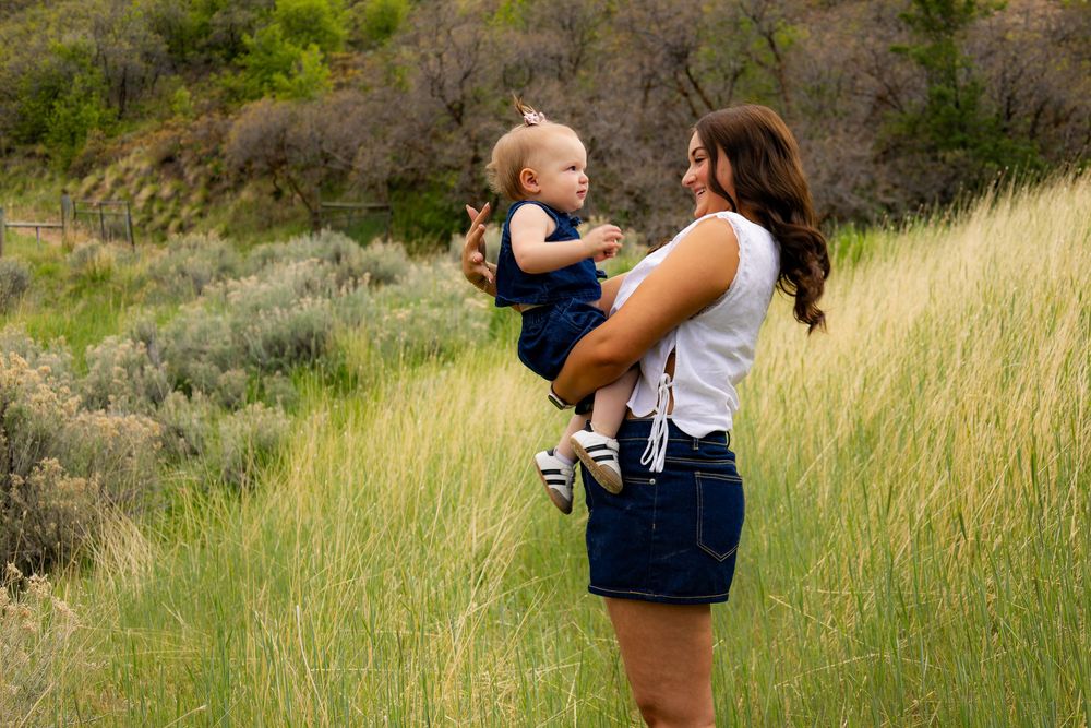 A mother holds her smiling baby up in a lush green meadow, both dressed in denim outfits on a sunny day.