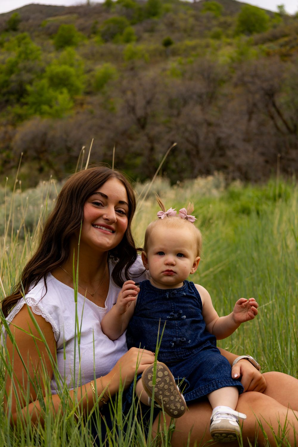 A smiling young woman sits in a green meadow holding a baby girl in a denim dress wearing a floral headband.