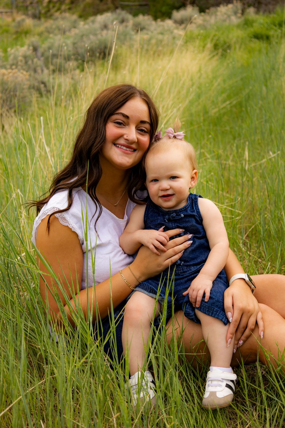 Young woman smiling with baby girl in denim romper sitting in tall green grass outdoors.
