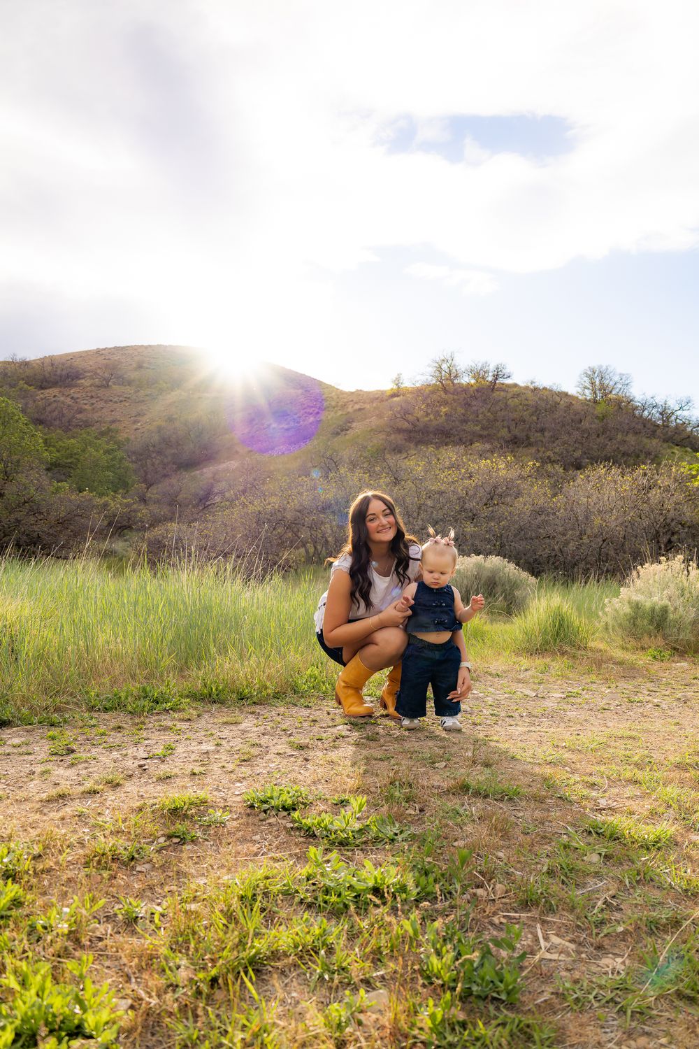 Mother crouching with toddler standing in sunny green meadow with hills in background.