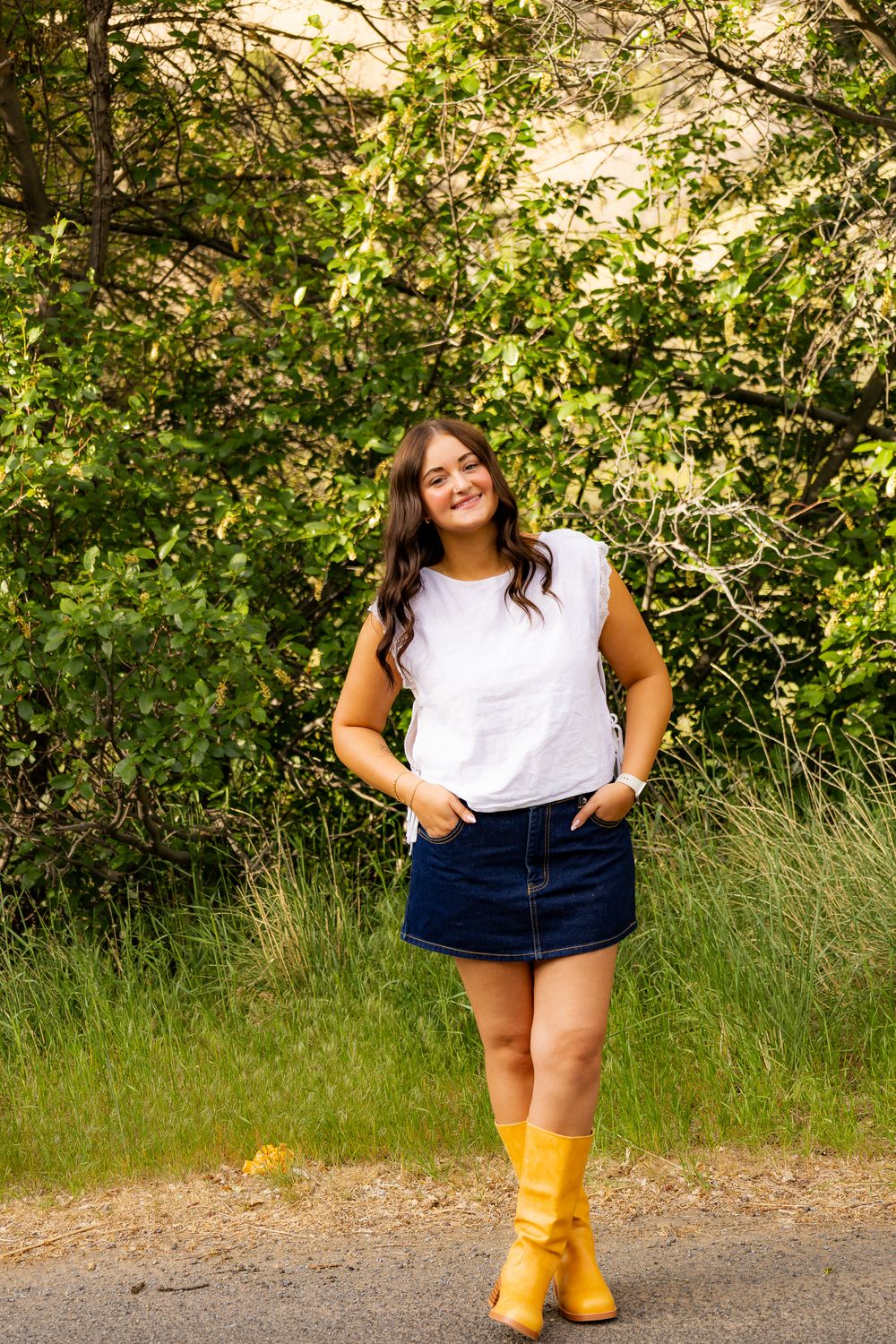 Brunette woman poses in white top, denim mini skirt, and yellow boots on a tree-lined path.