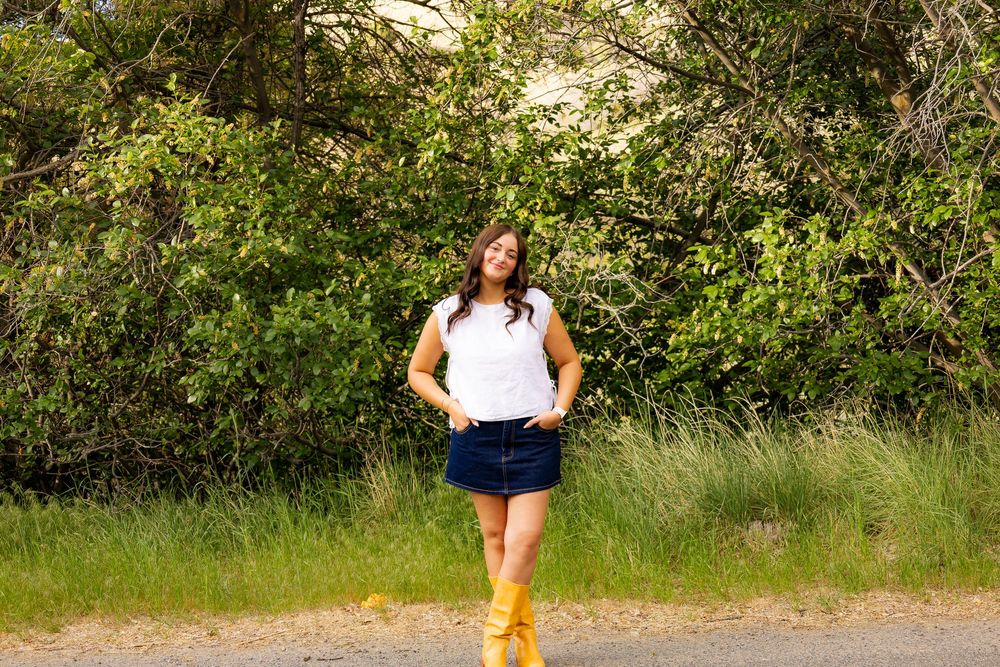 Woman in white sleeveless top, denim skirt, and yellow boots stands smiling on a rural road.