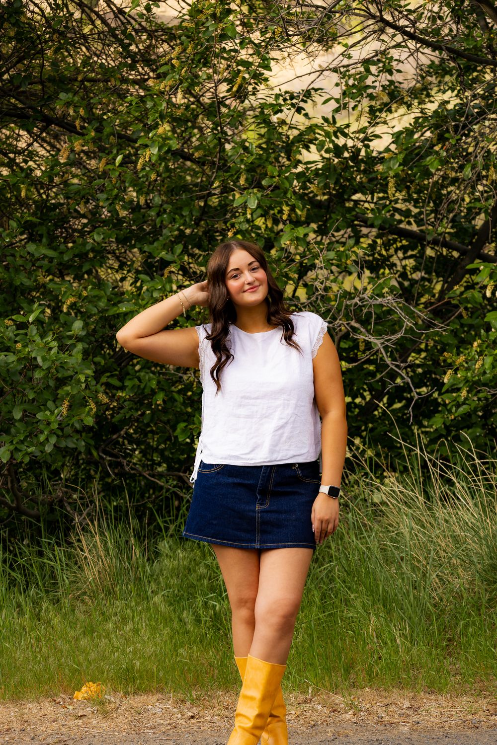 Smiling brunette woman in white top and denim skirt with yellow boots poses by lush green trees.