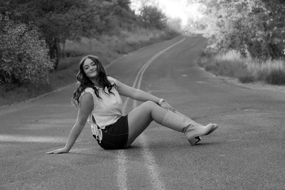 Black and white photo of a young woman sitting on a winding road, posing confidently with trees in the background.