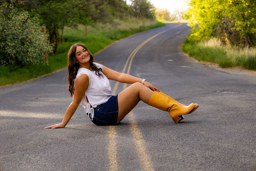 Young woman in white top, denim shorts, and yellow boots posing on a rural road.