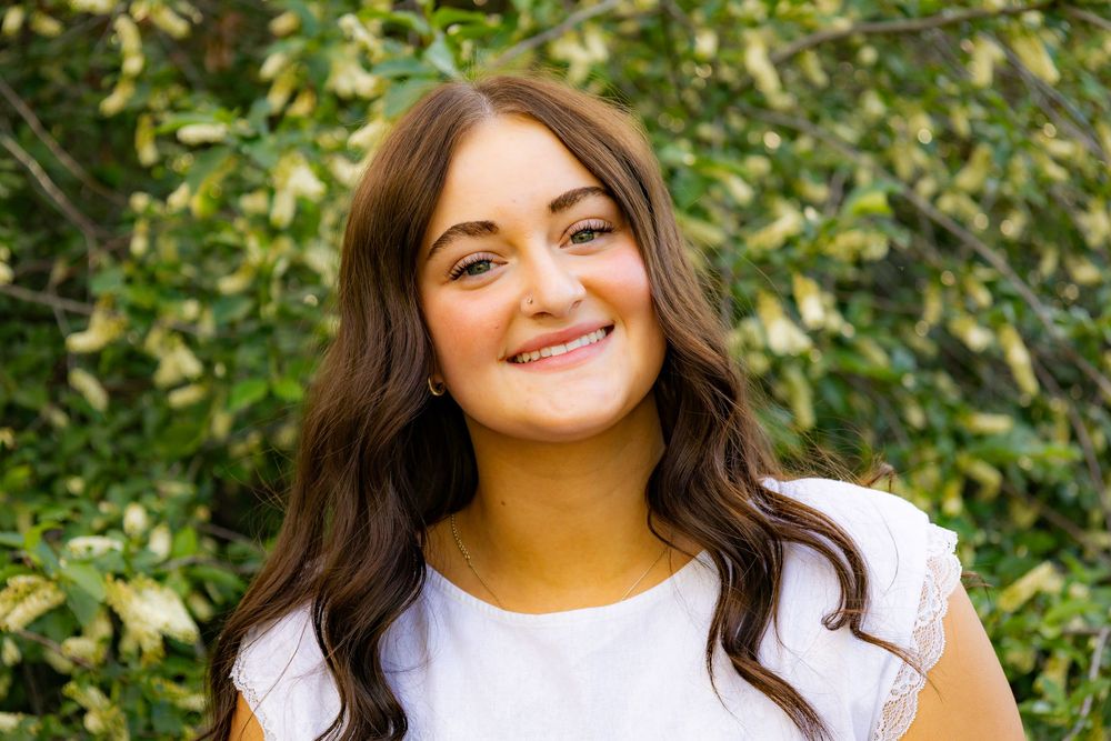 Young woman with long brown hair smiling outdoors, wearing a white top with green foliage background.