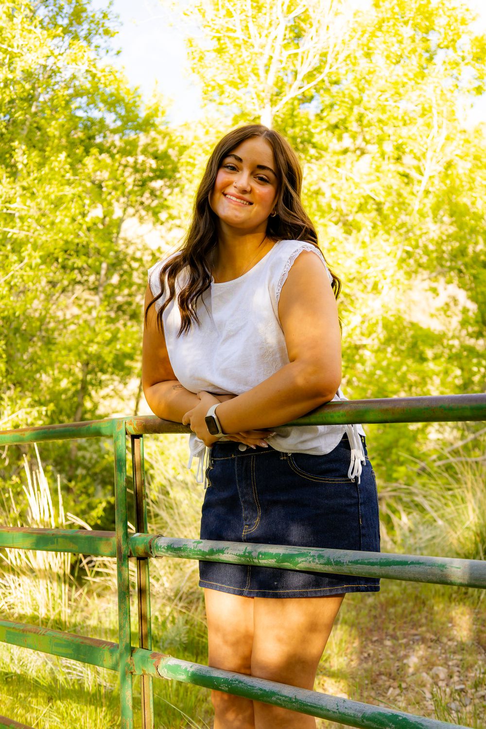 Young woman in white top and denim skirt smiling while leaning on a green metal fence outdoors.