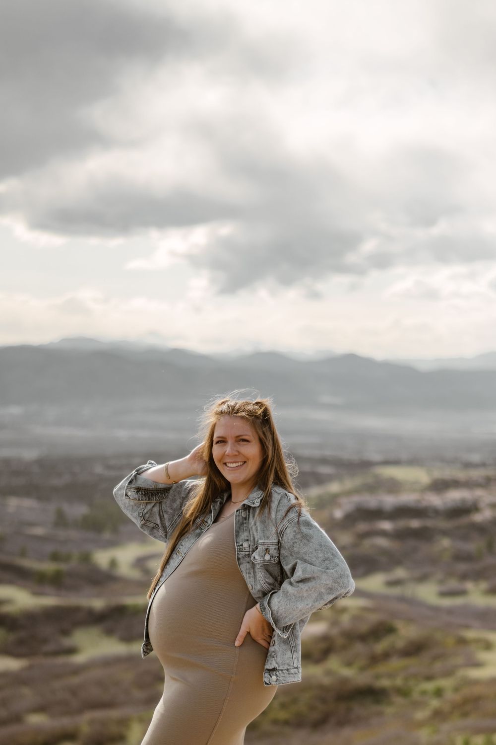 Woman in denim jacket poses confidently on hilltop with dramatic cloudy sky backdrop.