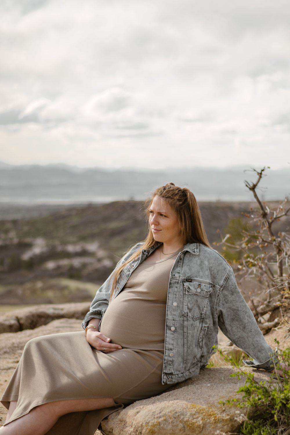 Expectant mother in brown dress and denim jacket reclines on rocky outcrop with mountain views.