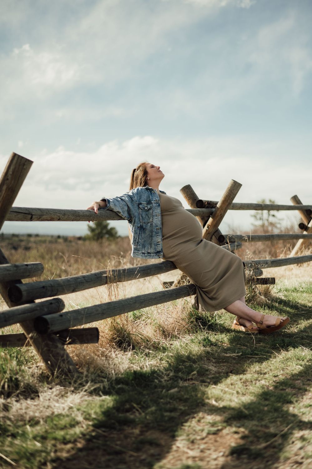 Person in denim jacket and dress lounges on wooden fence in natural setting.