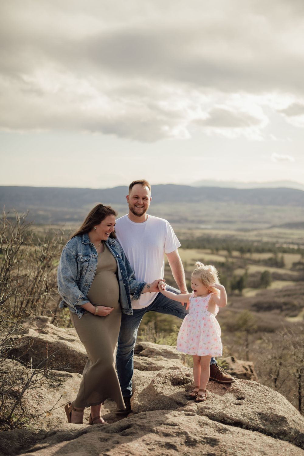 A couple and toddler share a tender moment on a rocky overlook with rolling hills in the background.