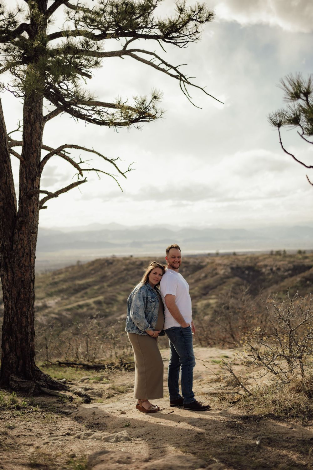 A couple sits together on a rocky hillside under moody skies and pine trees.