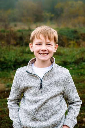 A child wearing a gray quarter-zip fleece sweater smiles outdoors against a blurred green natural background.