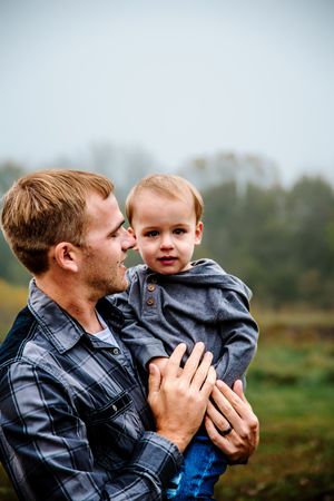A tender moment captured outdoors between a father in plaid shirt holding his young child against a misty natural backdrop.