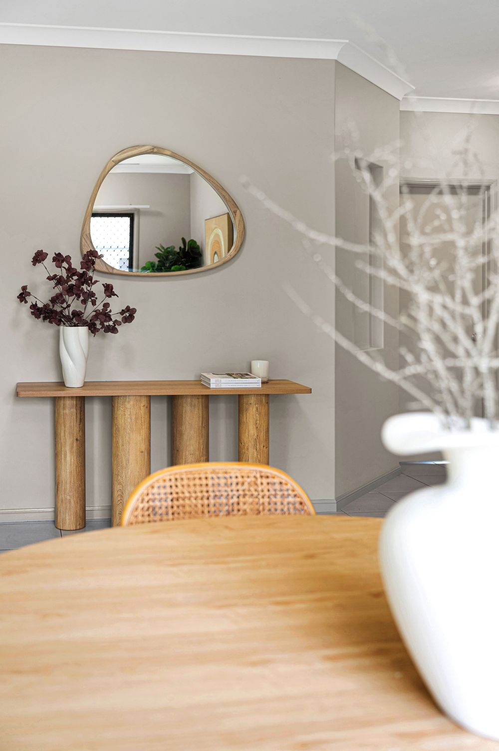 Modern minimalist dining area with wooden table and oval mirror on light gray walls.