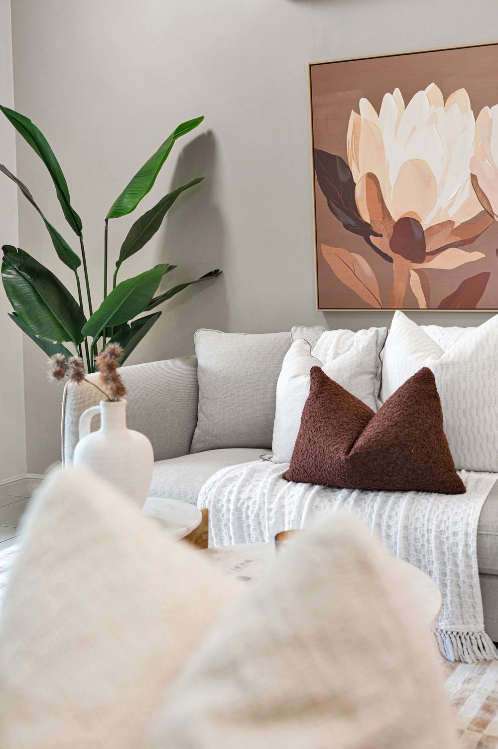 Minimalist living room decor featuring neutral tones, brown floral artwork, and tropical plants against a light gray wall.