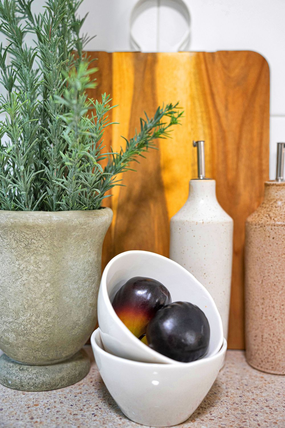 Kitchen counter with white ceramic bowls, dark plums and potted herbs against wooden background.
