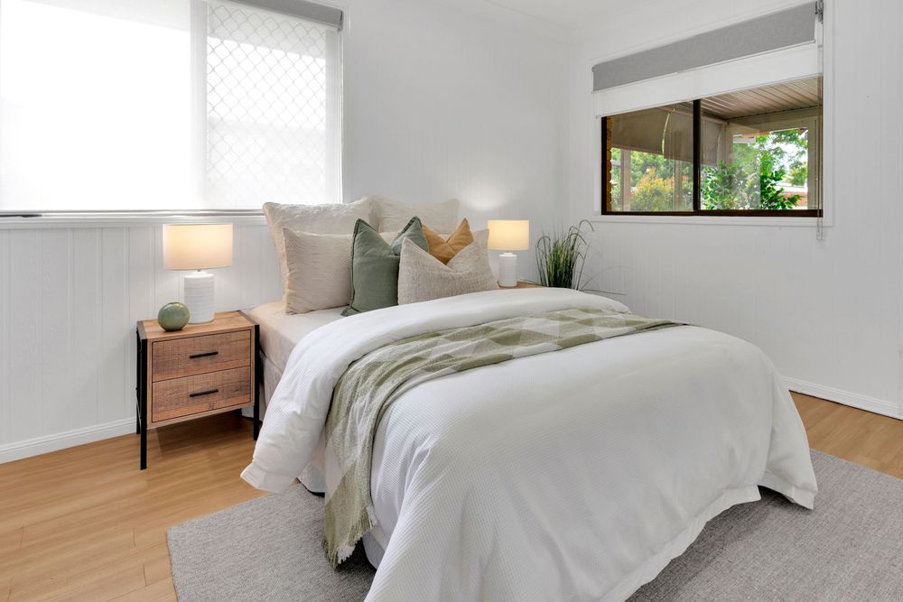Modern bedroom with white bedding, wooden nightstand, and green accents features natural light from two windows.