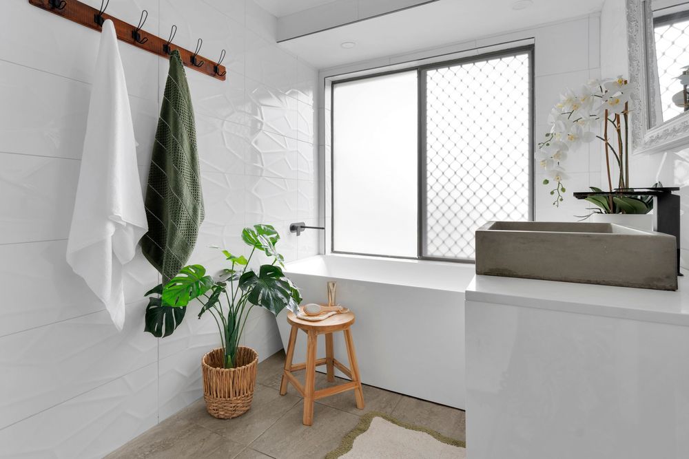 Modern white bathroom interior with natural light, plants, wooden stool and shower enclosure.