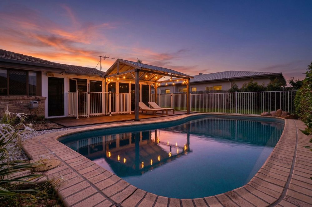 Swimming pool with wooden deck reflecting colorful sunset sky and luxury house in background at dusk.