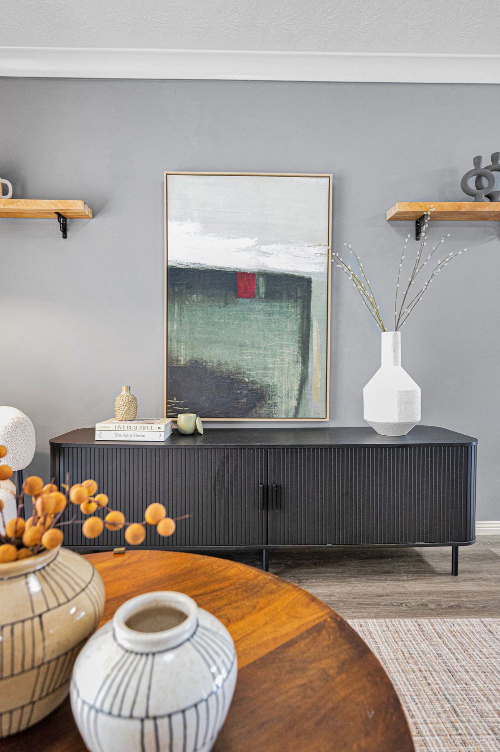 Modern living room features a black sideboard with orange pumpkins and white vases against a gray wall with artwork.