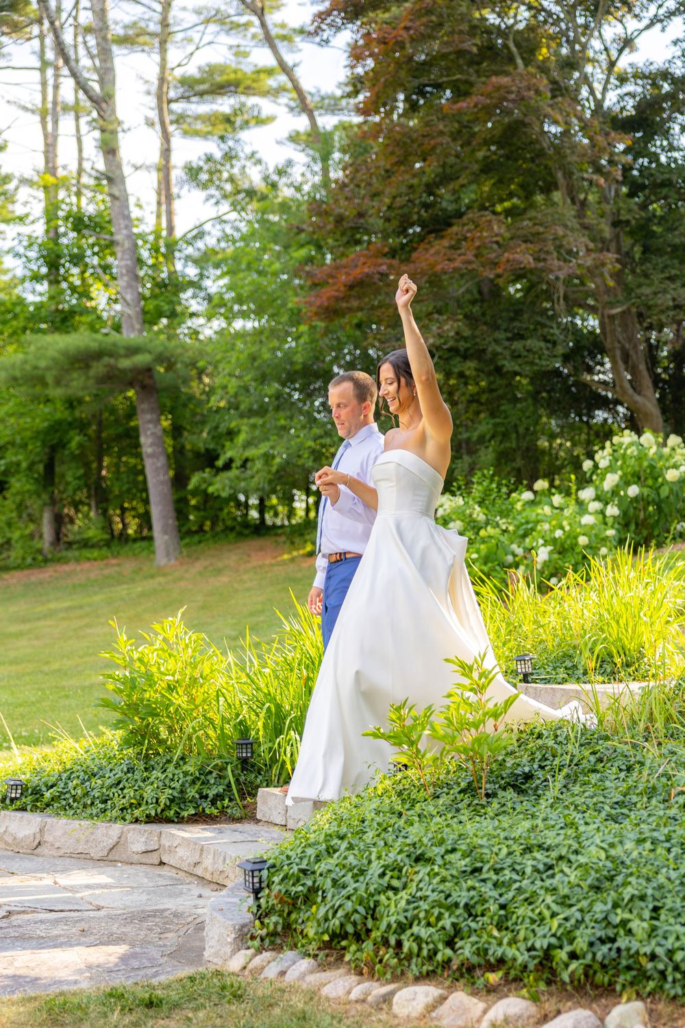 Bride in white gown raises arm joyfully as groom walks beside her through a lush garden wedding venue.