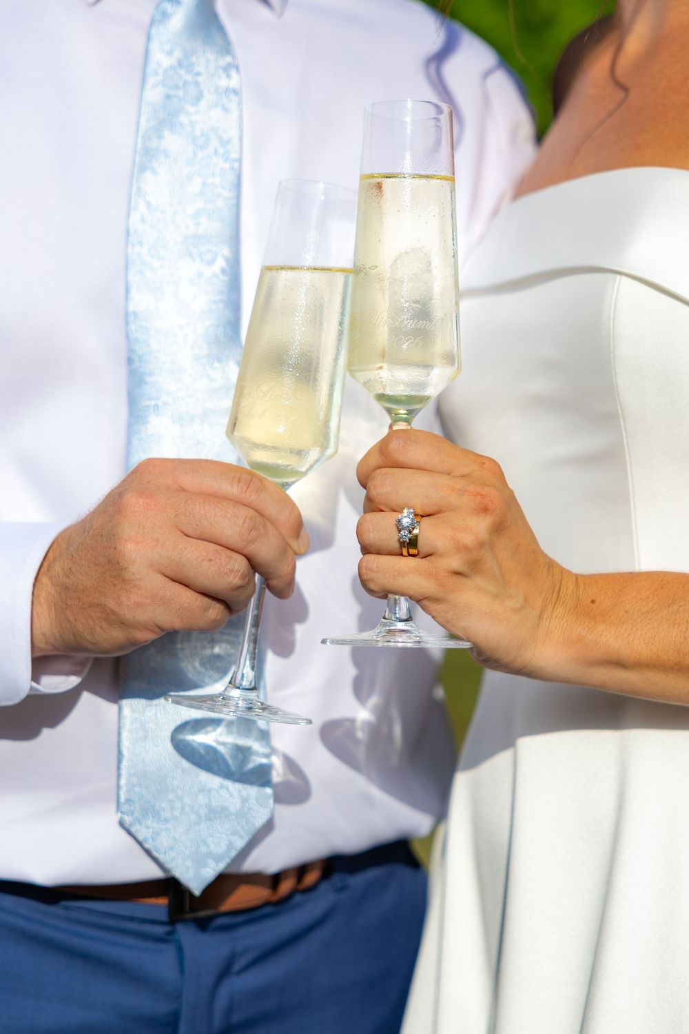 Newlywed couple toasting with champagne flutes, bride wearing diamond ring in white dress, groom in blue tie.