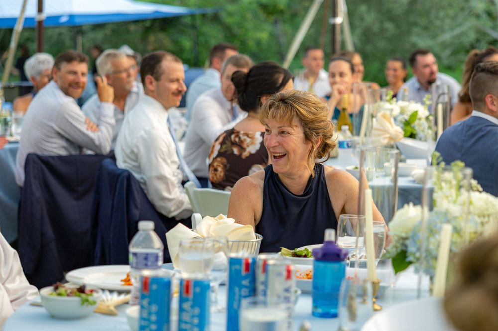 Guests laughing and enjoying a formal outdoor wedding reception dinner with elegant table settings and greenery.