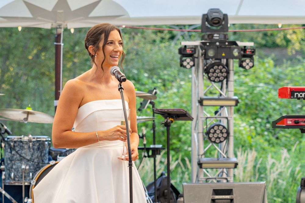 Woman in white strapless dress speaking at outdoor event microphone with stage lights and greenery background.
