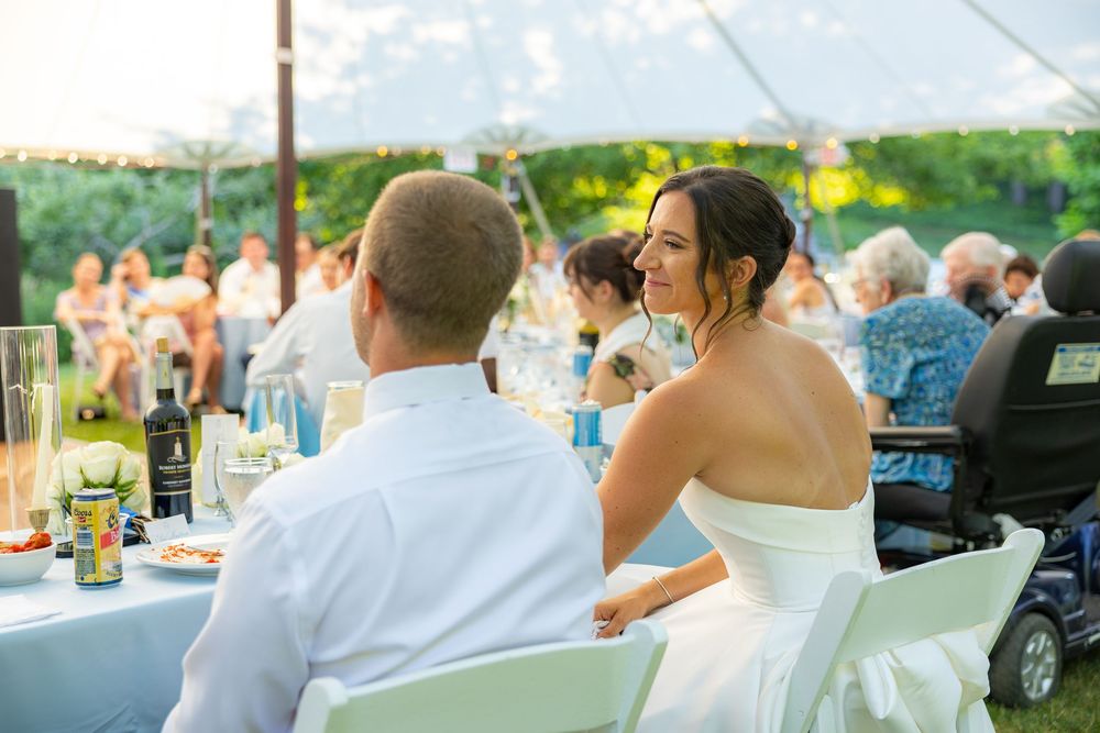 Bride in white strapless gown smiles at groom during outdoor wedding reception under a tent.