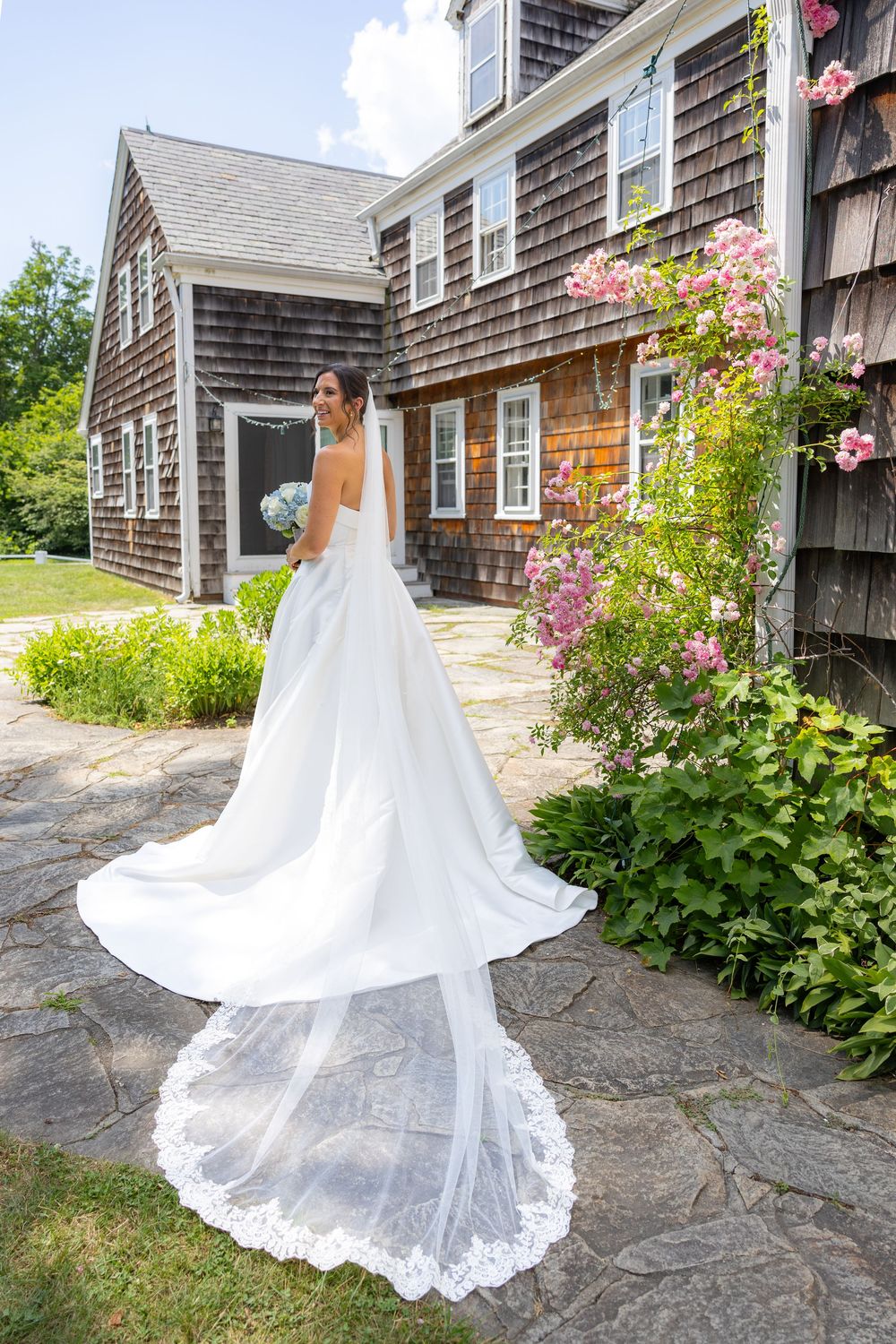 Bride in elegant white gown with lace cathedral veil stands on stone patio beside blooming roses at charming shingle-style home.