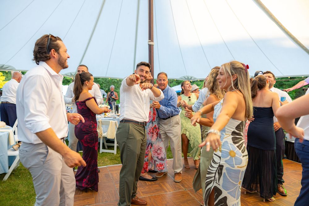 Guests dancing and celebrating under a white tent at an outdoor wedding reception on a sunny day.