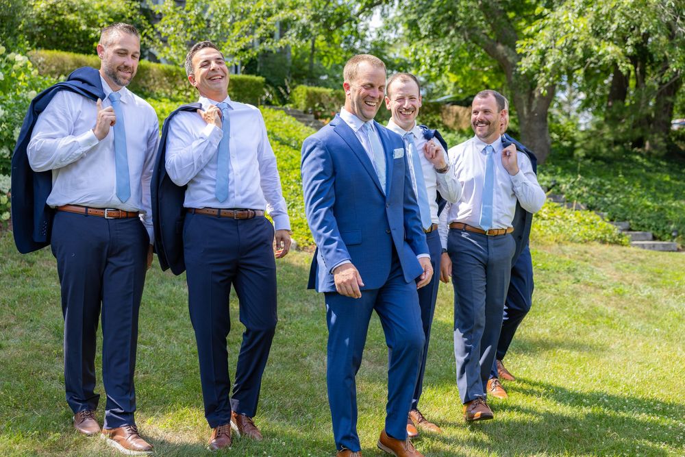 Five groomsmen in blue suits laughing and walking outdoors on a sunny day at a wedding.
